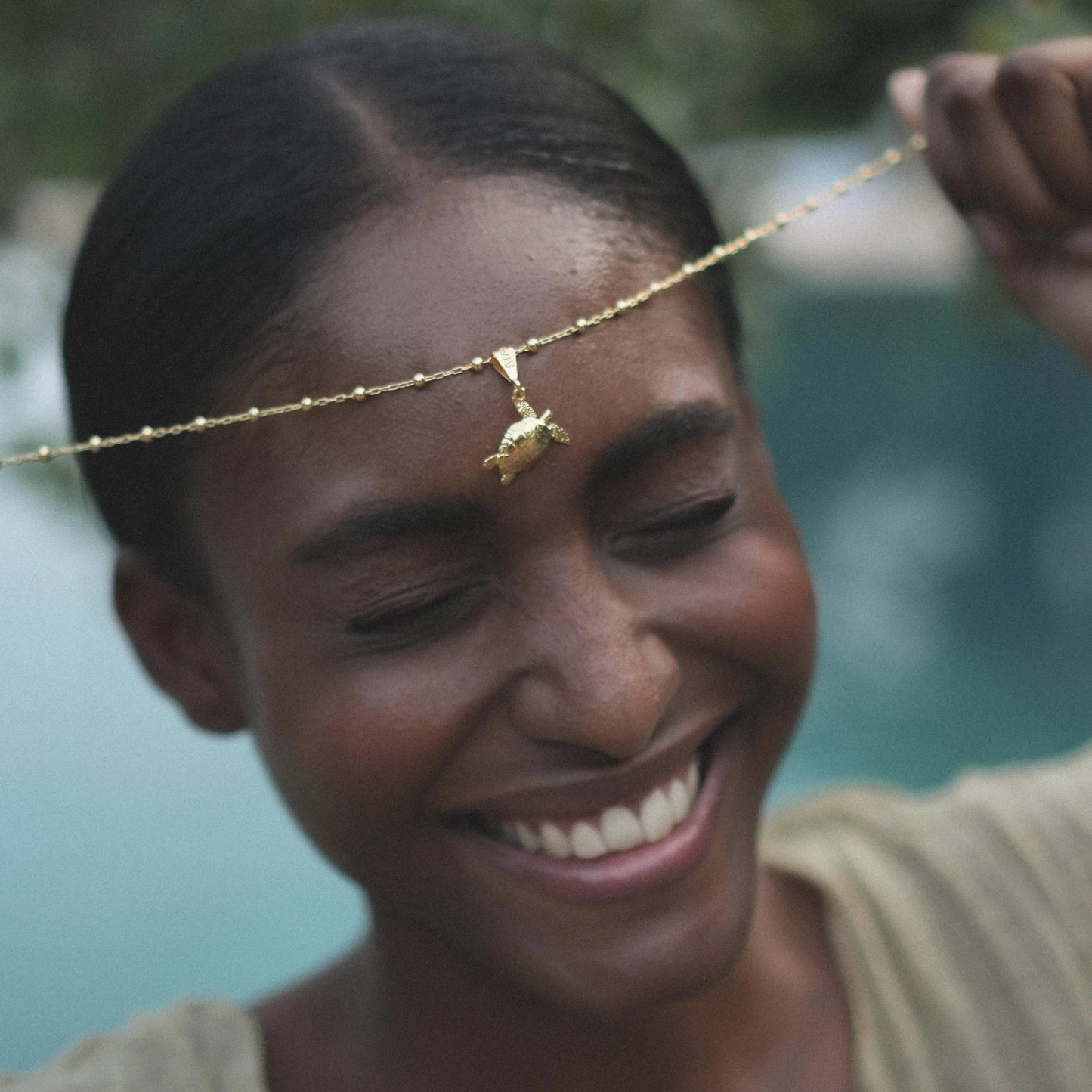 Modelo sonriendo con el colgante Cayume bañado en Oro con piscina turquesa de fondo.