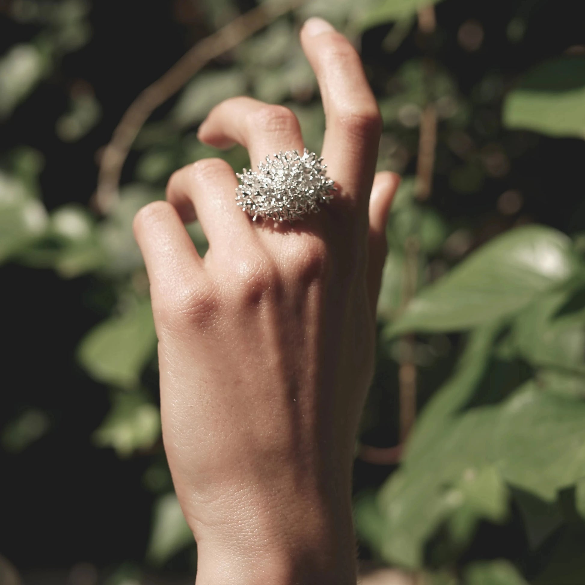 Detalles de la mano de una modelo luciendo el Anillo grande de Coralina en Plata con fondo de hojas verdes.