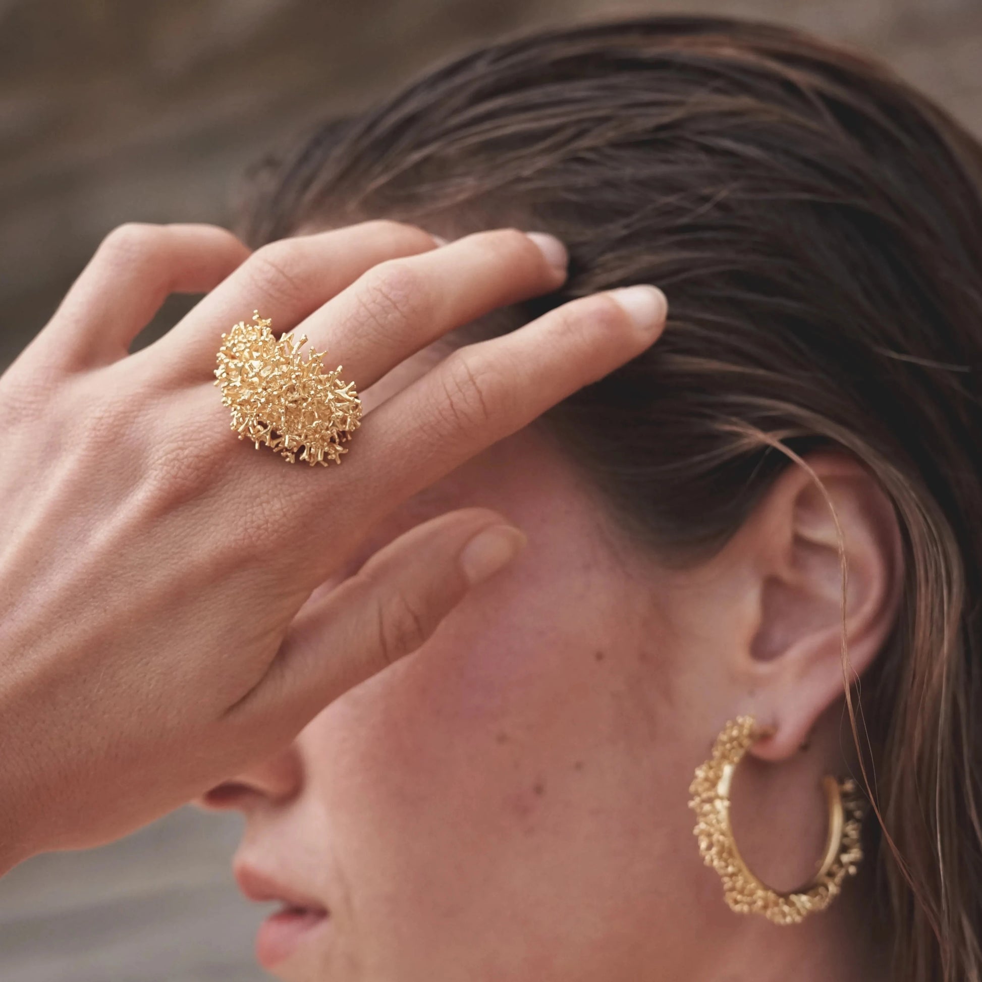 Modelo rozándose el pelo con la mano mientras luce el conjunto de joyas de la colección Coralina en Oro, en detalle el Anillo grande de Coralina bañado en Oro.