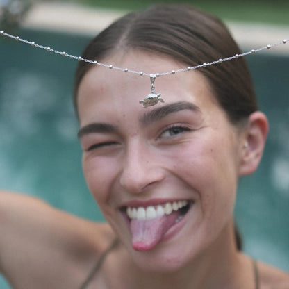 Modelo sacando la lengua y guiñando sujetando el Colgante de Cayume (Tortuga) en Plata con piscina color turquesa de fondo.